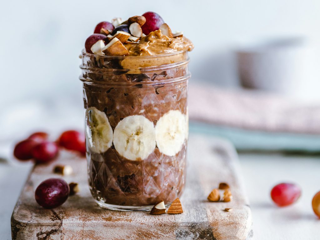 A Mason jar filled with creamy chocolate peanut butter banana overnight oats, garnished with sliced bananas, grapes, and chopped nuts. A spoon rests on the side, ready to scoop up the indulgent goodness.