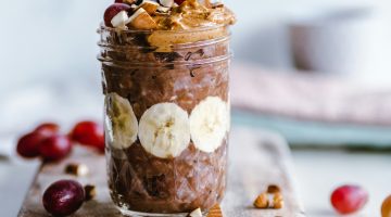 A Mason jar filled with creamy chocolate peanut butter banana overnight oats, garnished with sliced bananas, grapes, and chopped nuts. A spoon rests on the side, ready to scoop up the indulgent goodness.