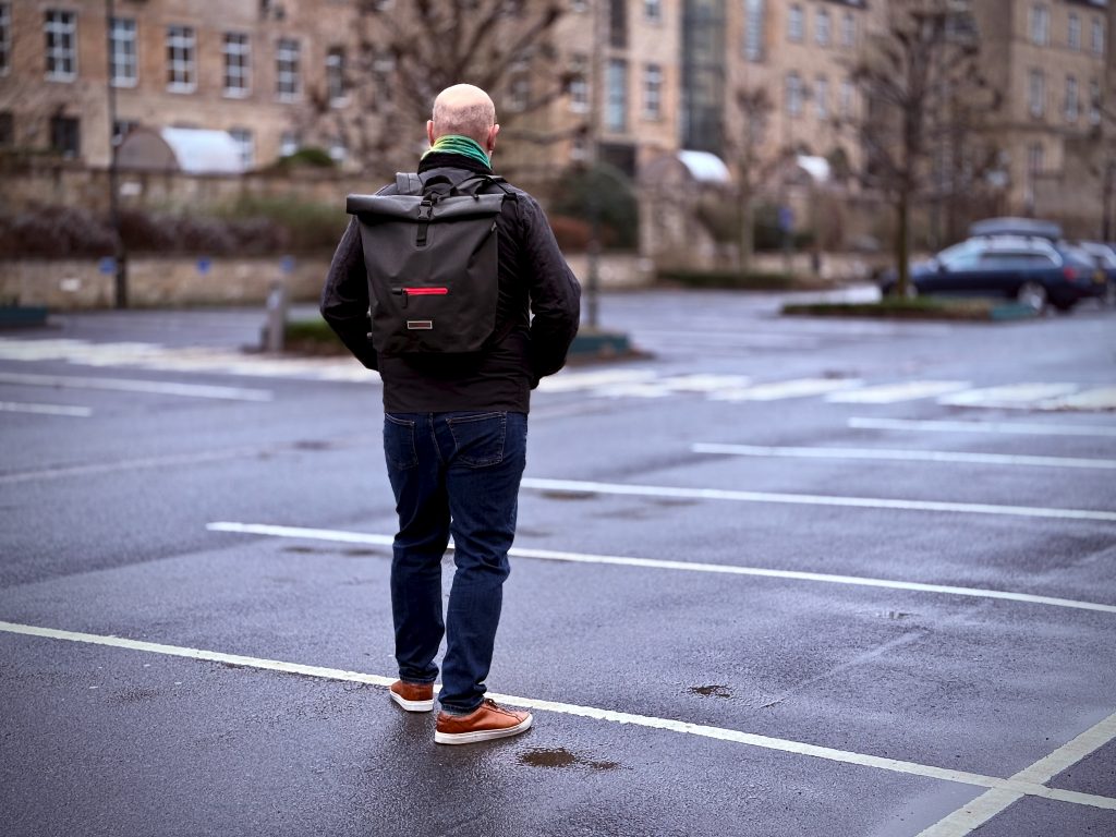 A man stands in an empty, wet parking lot, facing away from the camera. He is wearing a black jacket, dark jeans, brown shoes, and a stylish black roll-top backpack with red accents. The scene is set in front of an elegant, historic stone building with rows of windows, and a few cars are visible in the distance. The atmosphere is overcast, with a damp, reflective ground suggesting recent rain.