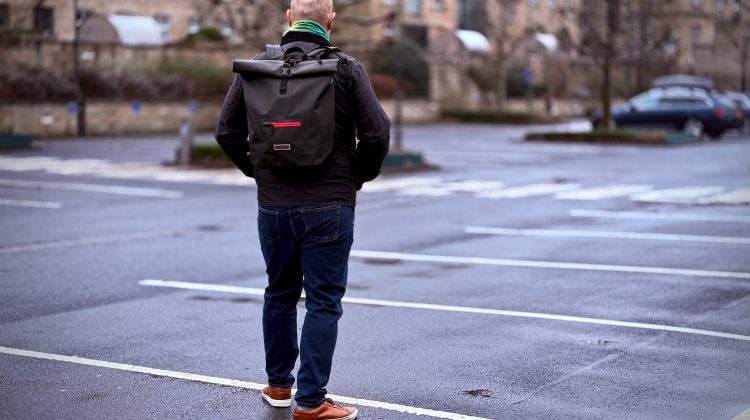 A man stands in an empty, wet parking lot, facing away from the camera. He is wearing a black jacket, dark jeans, brown shoes, and a stylish black roll-top backpack with red accents. The scene is set in front of an elegant, historic stone building with rows of windows, and a few cars are visible in the distance. The atmosphere is overcast, with a damp, reflective ground suggesting recent rain.