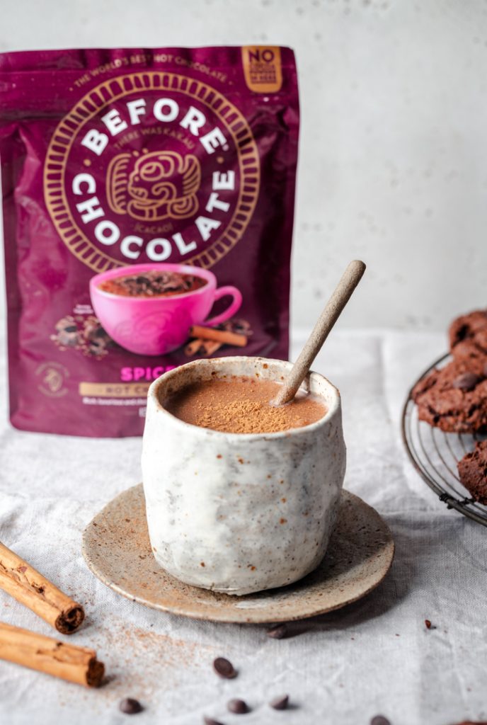 A mug of rich, creamy hot chocolate topped with cinnamon, placed on a rustic saucer. In the background, a vibrant packet of "Before Chocolate" hot chocolate mix with the tagline "No Refined Sugar" is visible, along with cinnamon sticks and chocolate cookies on a cooling rack, creating a warm and inviting scene.