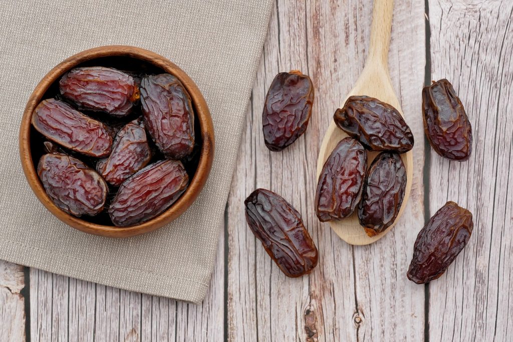 Wooden bowl filled with Medjool dates on a rustic wooden table, accompanied by additional dates on a wooden spoon and cloth napkin.