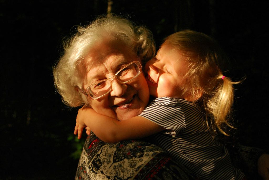 An elderly woman with glasses smiling warmly while being hugged by a young girl with blonde hair in a striped shirt, surrounded by soft natural lighting.