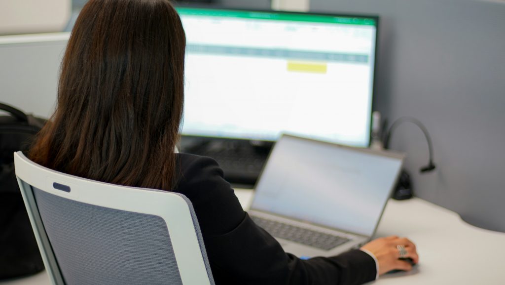 A woman working on a computer at an office desk, focusing on a spreadsheet displayed on a large monitor.