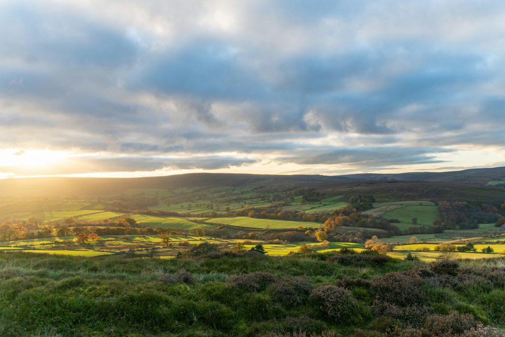 Scenic view of the Yorkshire countryside at sunset, featuring rolling green hills, fields, and a dramatic cloudy sky illuminated by golden sunlight.