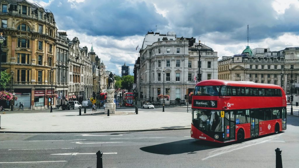 London street scene featuring a red double-decker bus on route 15 to Blackwall, surrounded by historic architecture, with a cloudy sky overhead.