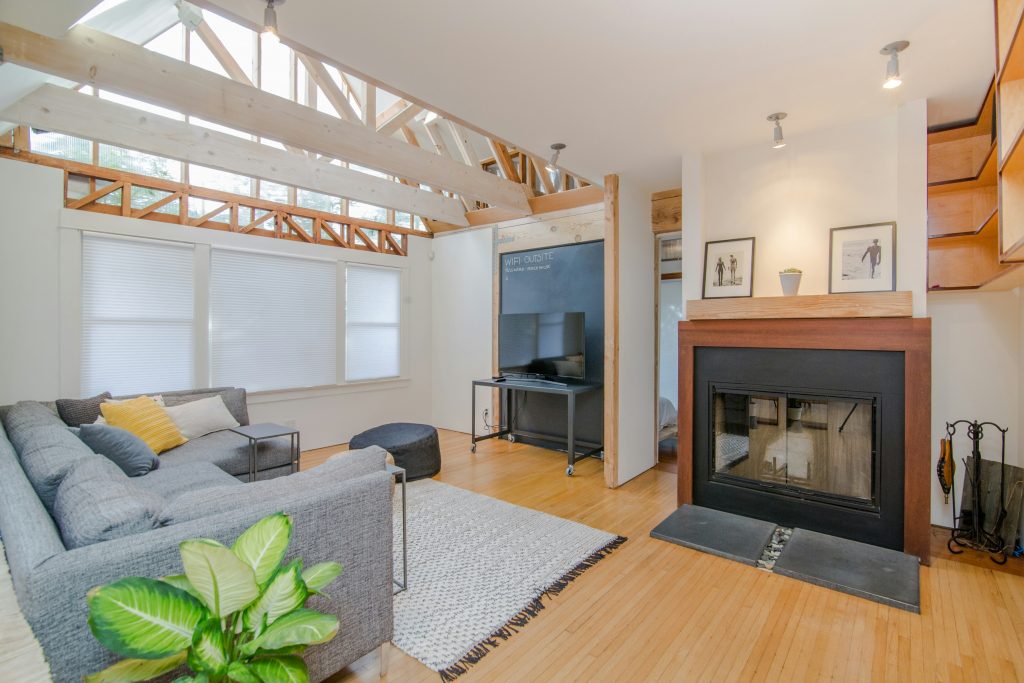 Modern living room interior featuring exposed wooden beams, a cozy gray sectional sofa, a fireplace with wooden accents, and bright natural lighting from large windows.