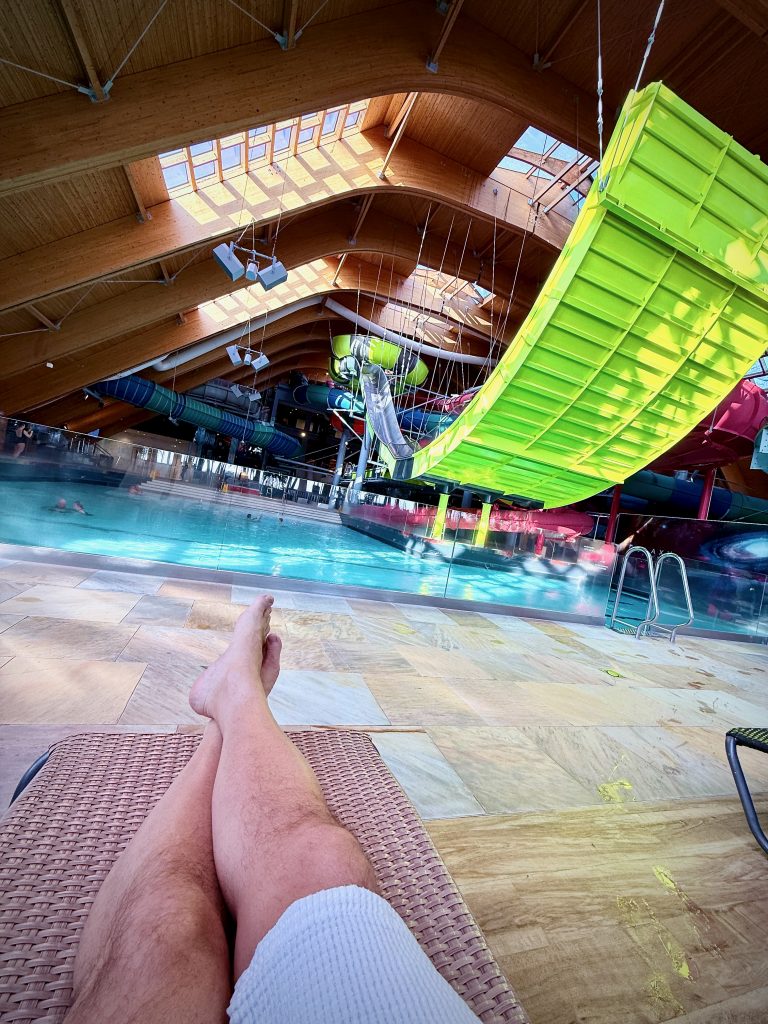 A person relaxing on a sun lounger at Therme Bucharest, overlooking an indoor pool and a bright yellow water slide.