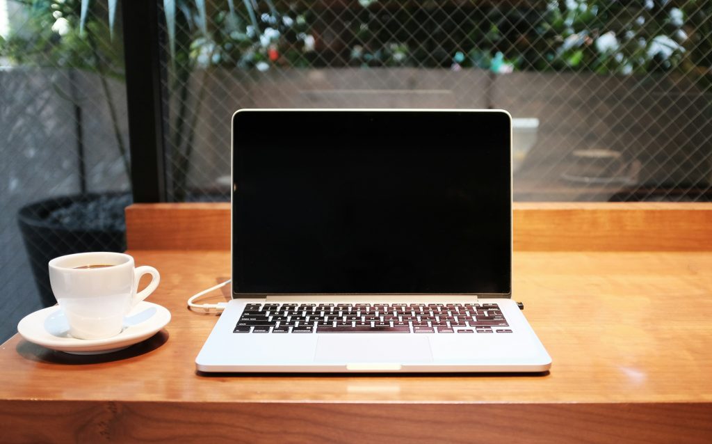 A laptop with a black screen sits on a wooden table next to a white coffee cup and saucer. The setting appears to be a café with a wire-mesh window and greenery in the background.