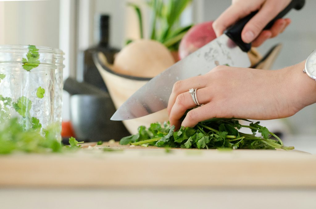 A close-up of a person chopping fresh cilantro on a wooden cutting board with a sharp knife. The person is wearing a silver watch and a diamond ring, and the background features a kitchen setting with fresh produce and a glass jar.