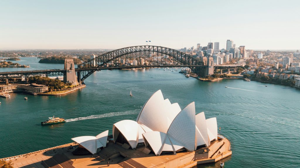 A stunning aerial view of Sydney, Australia, featuring the iconic Sydney Opera House in the foreground and the Sydney Harbour Bridge spanning across the harbour. The city's skyline and surrounding waters are illuminated by golden sunlight, with boats cruising through the harbour.