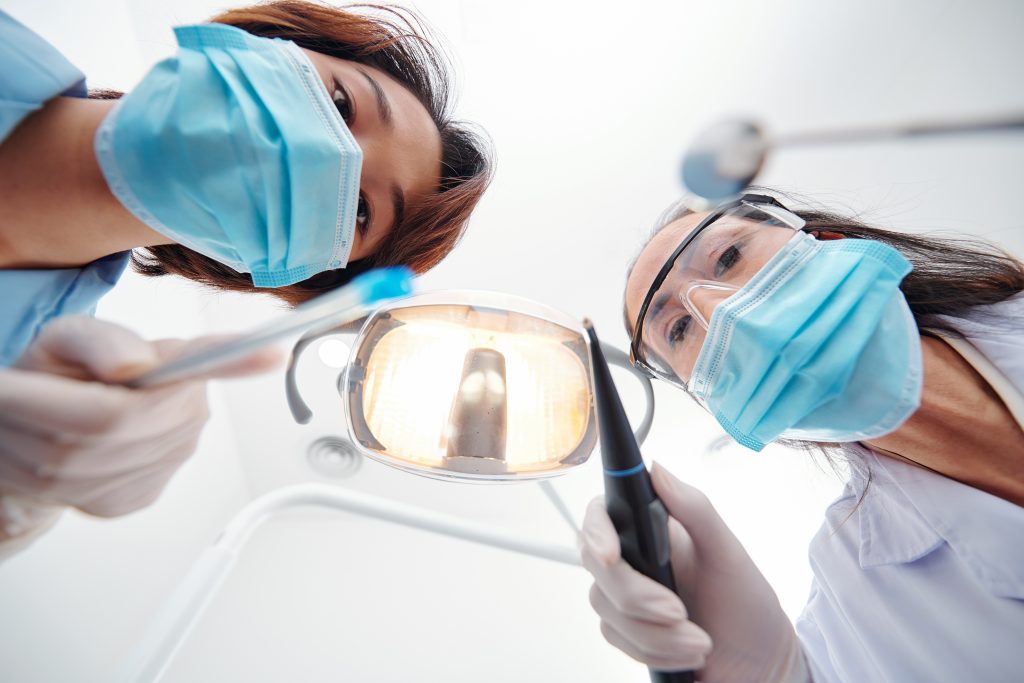 Two dentists wearing face masks and gloves are examining a patient from a close-up, first-person perspective. One holds a dental mirror, while the other holds a dental tool under a bright examination light.