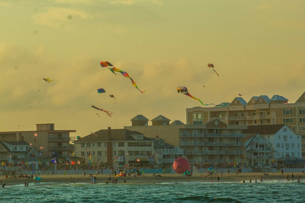 A vibrant beach scene at sunset in Ocean City, Maryland, with colorful kites soaring in the sky above beachfront buildings and a lively crowd enjoying the shoreline.