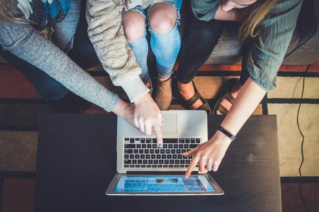 Three people sitting closely together on a couch, pointing at the screen of an open laptop placed on a black table. The image captures a top-down perspective, showing their hands actively engaging with the device, suggesting collaboration or excitement over something on the screen.