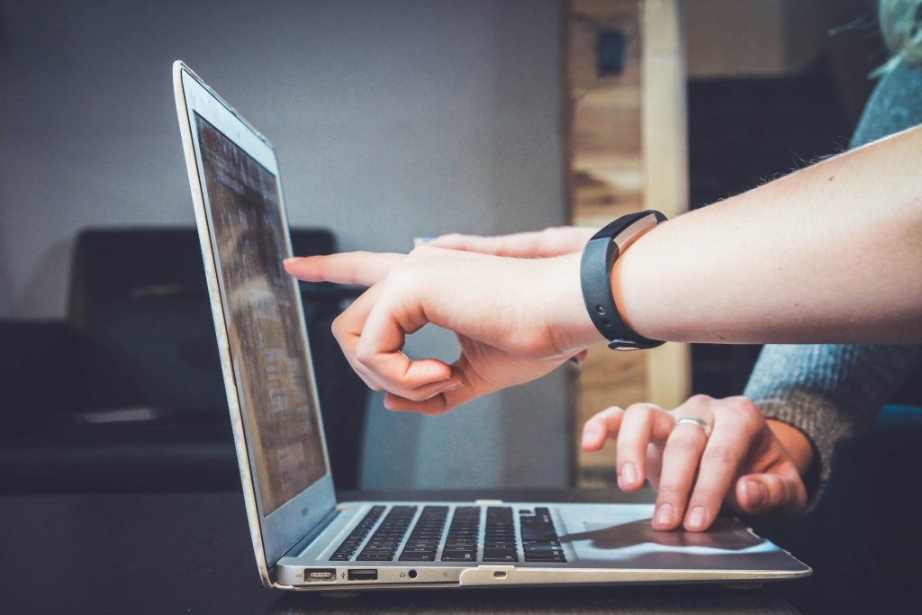 Close-up of two people using a laptop, with one person pointing at the screen while the other types. The setting appears to be an indoor workspace, with a blurred background.