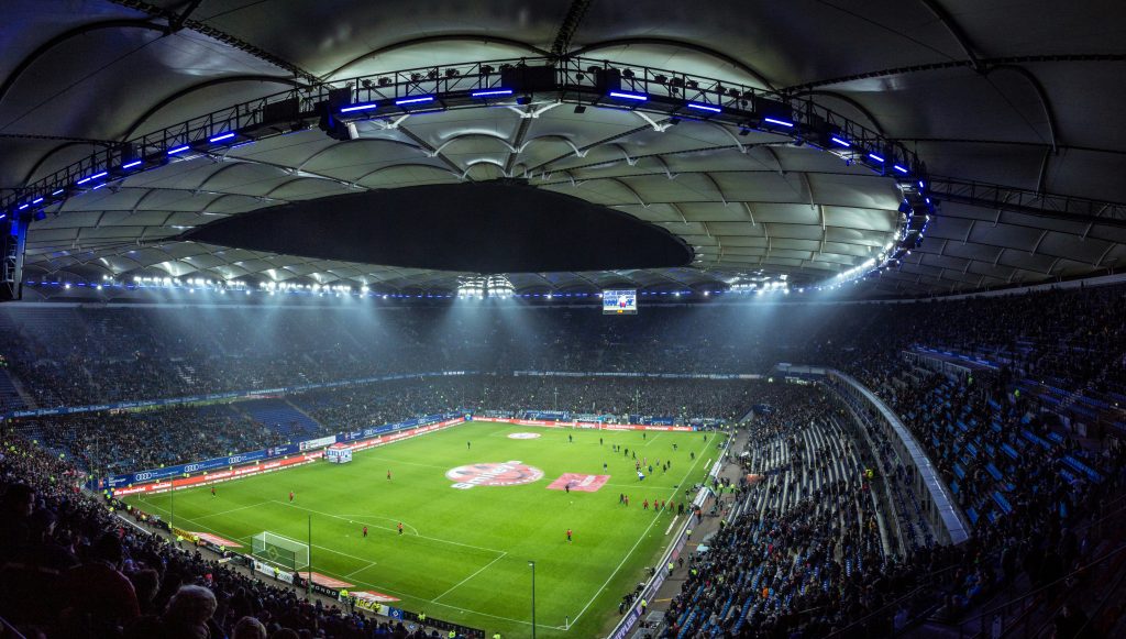 Panoramic view of a brightly lit football stadium at night, filled with spectators, as players warm up on the field before a match.