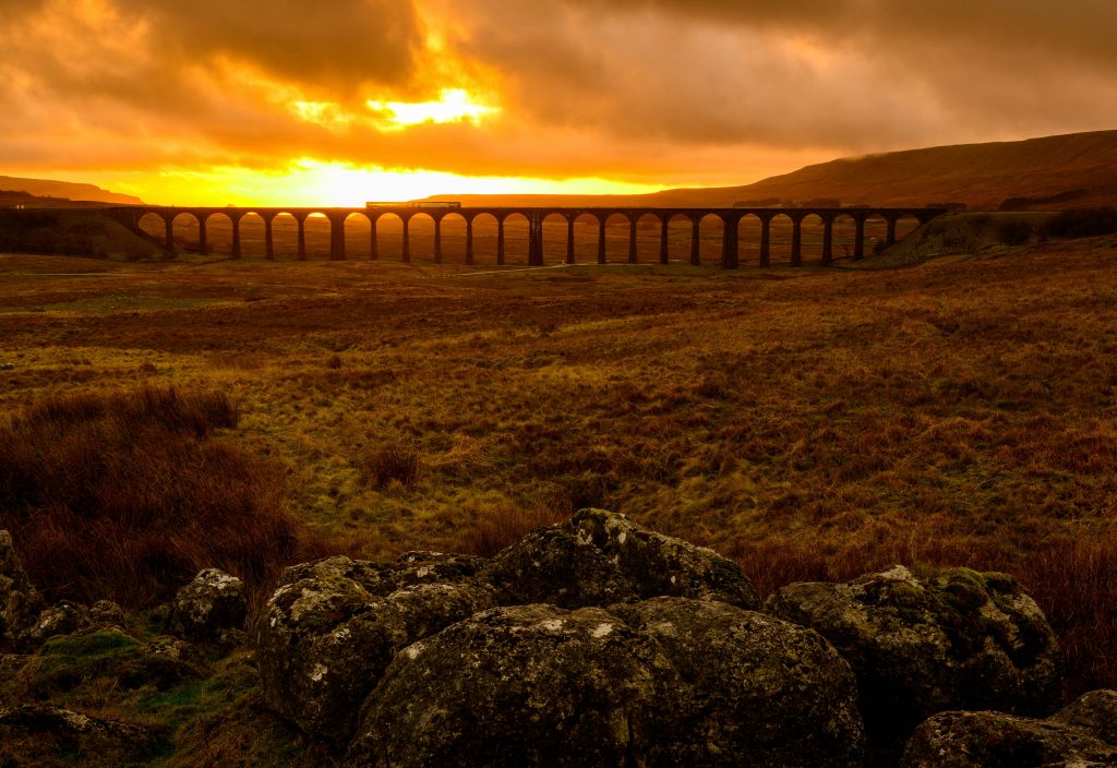 A stunning view of a viaduct stretching across the Yorkshire countryside at sunset, with golden hues illuminating the sky and rolling hills in the background.
