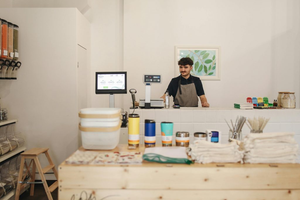A friendly shopkeeper wearing an apron stands behind the counter of a sustainable health and wellness store. The store features bulk food dispensers, eco-friendly products, reusable containers, and natural health items, creating an inviting and environmentally conscious shopping atmosphere.