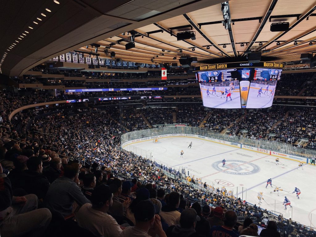 A packed Madison Square Garden during an NHL game, with fans filling the arena and players from two teams engaged in action on the ice. The large overhead scoreboard displays live game footage and scores, enhancing the vibrant atmosphere of the game night.