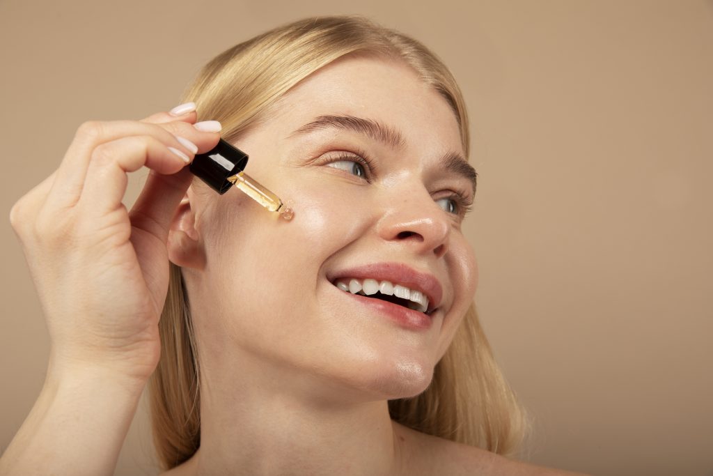 A smiling blonde woman with clear, glowing skin applies a drop of facial serum to her cheek using a glass dropper. The background is a warm, neutral tone, enhancing the image's beauty and skincare theme