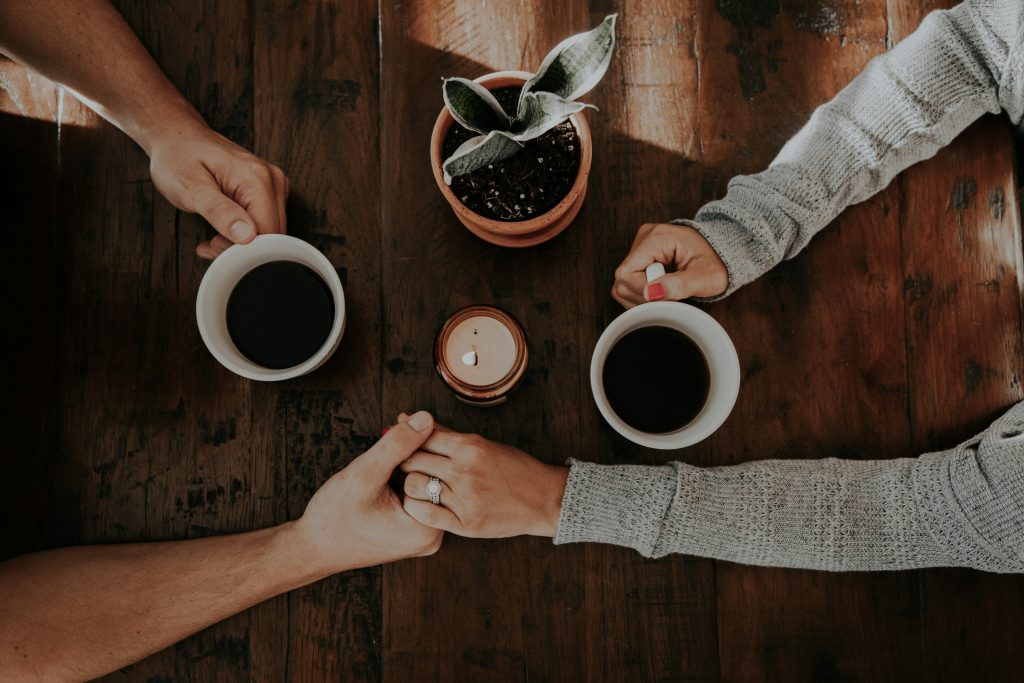 A romantic top-down view of a couple holding hands across a rustic wooden table, each holding a cup of black coffee. A small candle and a potted plant sit between them, adding to the cosy and intimate atmosphere. One person wears a knitted sweater, and the other has a visible engagement ring, suggesting warmth, love, and connection.
