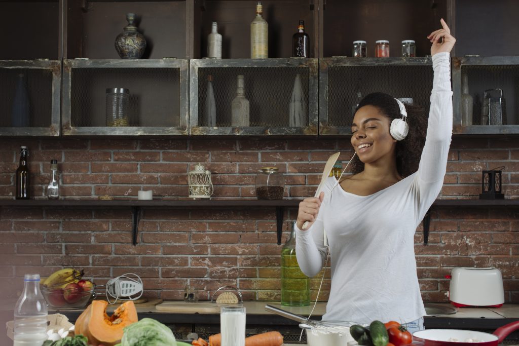 A young woman wearing white headphones and a long-sleeved shirt dances joyfully in the kitchen while holding a wooden spoon as a microphone. The rustic kitchen setting features a brick wall, open shelves with bottles and jars, and fresh ingredients like vegetables and milk on the countertop