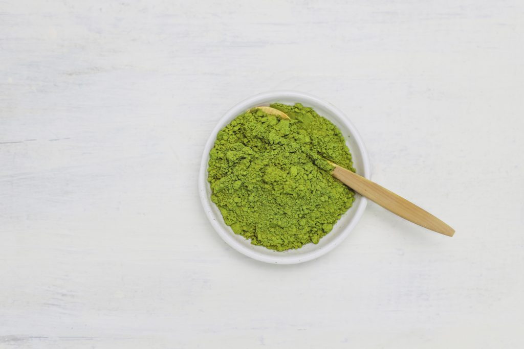 A small white bowl filled with vibrant green matcha powder, accompanied by a wooden spoon, set on a white textured surface.