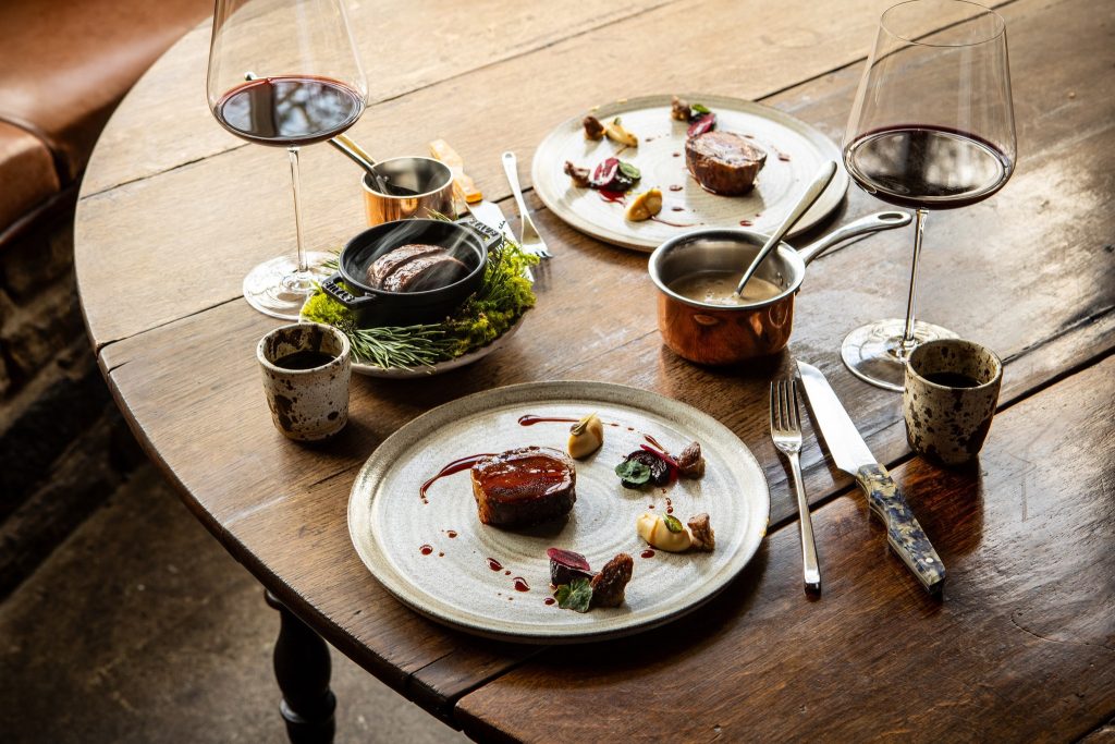 A beautifully composed dish featuring succulent cuts of meat, served with a rich sauce, delicate vegetable garnishes, and a side of creamy purée. The rustic wooden table setting includes copper cookware, elegant wine glasses, and speckled ceramic cups, creating an inviting and luxurious dining atmosphere.