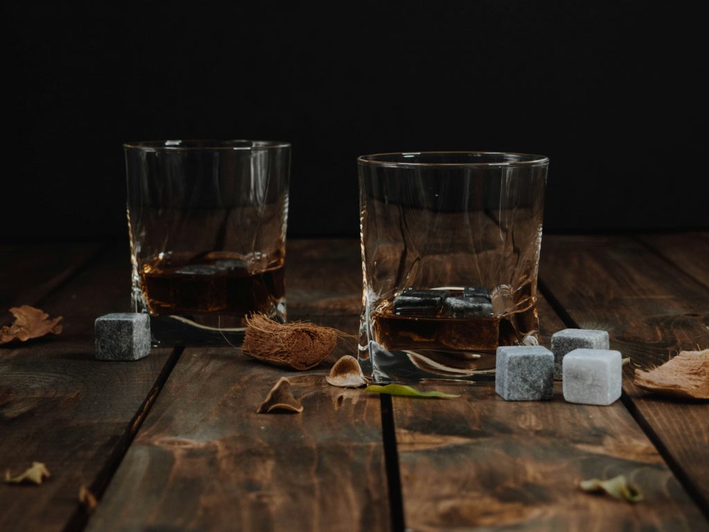 Two glasses of whisky on a rustic wooden table, accompanied by whisky stones and dried botanicals, creating a warm and sophisticated atmosphere.