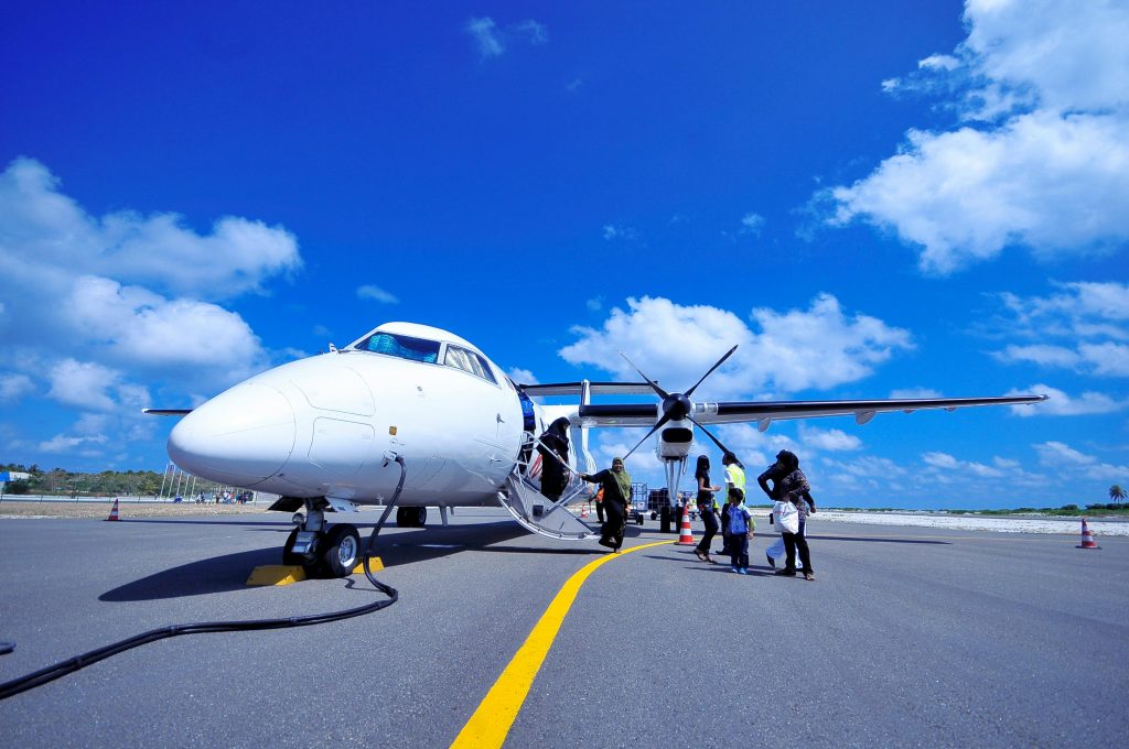 A white private jet with a propeller stands on the tarmac under a bright blue sky with scattered clouds. A group of passengers, including women and children, are seen boarding the aircraft via a small staircase. A ground crew member in a high-visibility vest assists them. The setting appears to be a tropical or remote airport, with palm trees visible in the background.