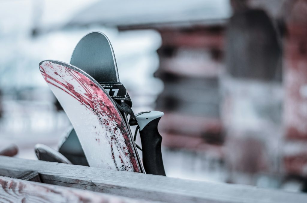 Close-up of a pair of skis with red and white markings resting against a wooden rack, with a blurred snowy mountain lodge in the background.