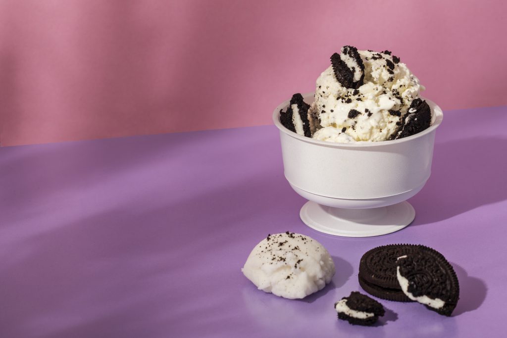 A bowl of cookies and cream ice cream with crushed chocolate sandwich cookies, placed on a pastel pink and purple background, with extra cookie pieces on the surface.