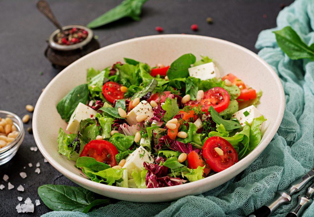 A fresh and vibrant garden salad in a ceramic bowl, featuring cherry tomatoes, feta cheese, mixed greens, spinach, and pine nuts, garnished with herbs and drizzled with dressing. The dish is set on a rustic dark surface with scattered salt, a small bowl of pine nuts, and a textured blue cloth in the background.