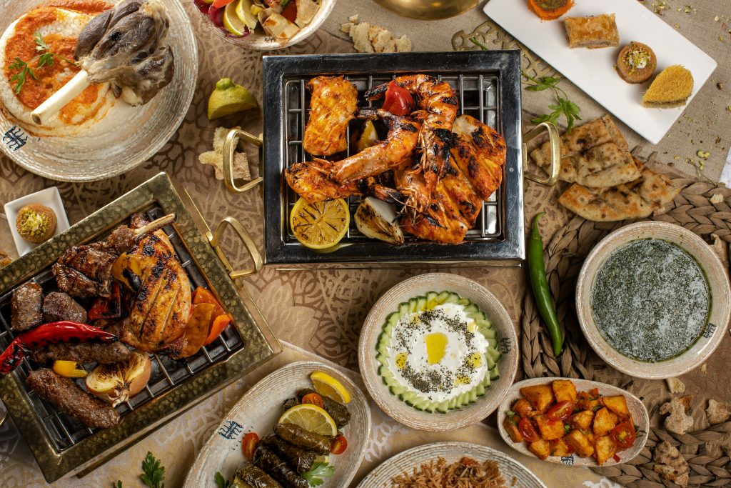 A vibrant spread of Middle Eastern cuisine displayed on an ornate table setting. The feast includes grilled meats, seafood, lamb shank over rice, stuffed grape leaves, creamy yogurt dip, flatbread, spiced potatoes, and a selection of traditional desserts. The dishes are arranged in decorative plates and trays, showcasing rich textures and warm, inviting colors.