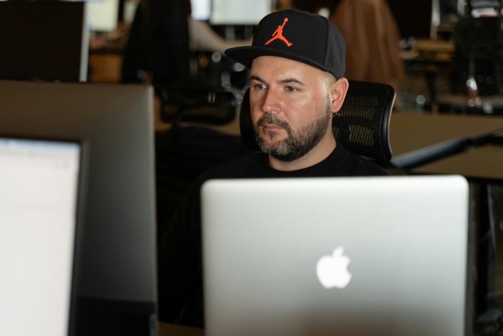 A bearded man wearing a black cap with an orange Jumpman logo sits at a desk, focused on his laptop screen. He appears concentrated and serious, working in a dimly lit office environment with multiple computer monitors in the background.