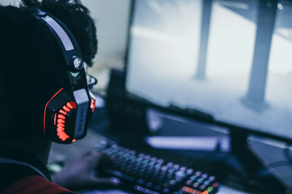 A gamer wearing a headset with red LED lights is immersed in playing a video game on a computer screen. The blurred screen displays a foggy, atmospheric scene, while a mechanical keyboard with RGB lighting is visible in the foreground.