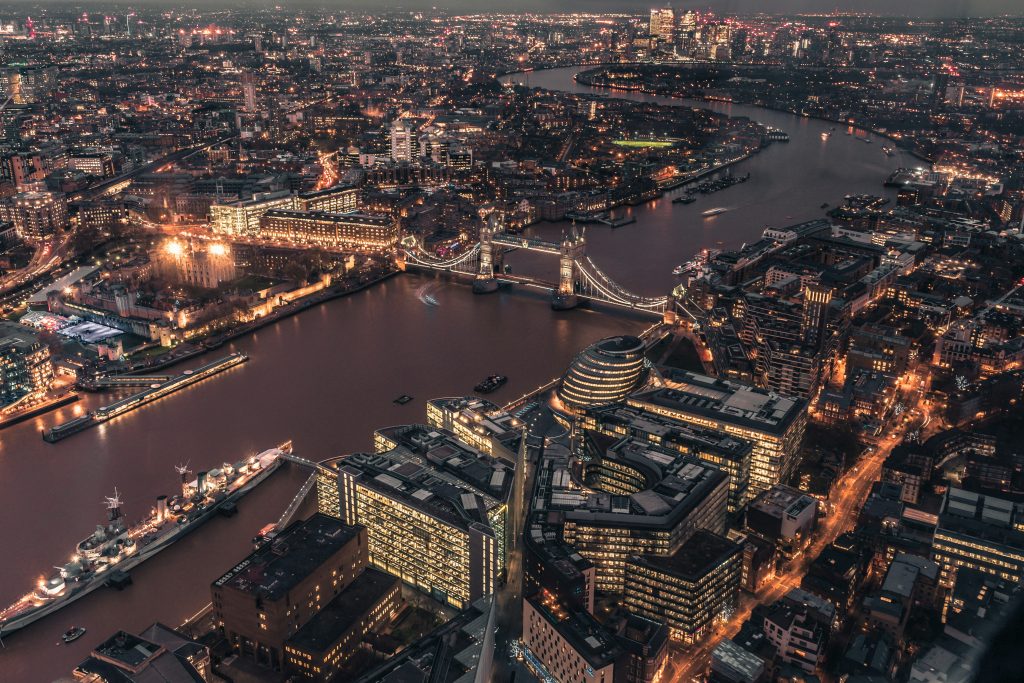 Aerial night view of London featuring the illuminated Tower Bridge, the River Thames, and surrounding city lights, including historic and modern buildings.