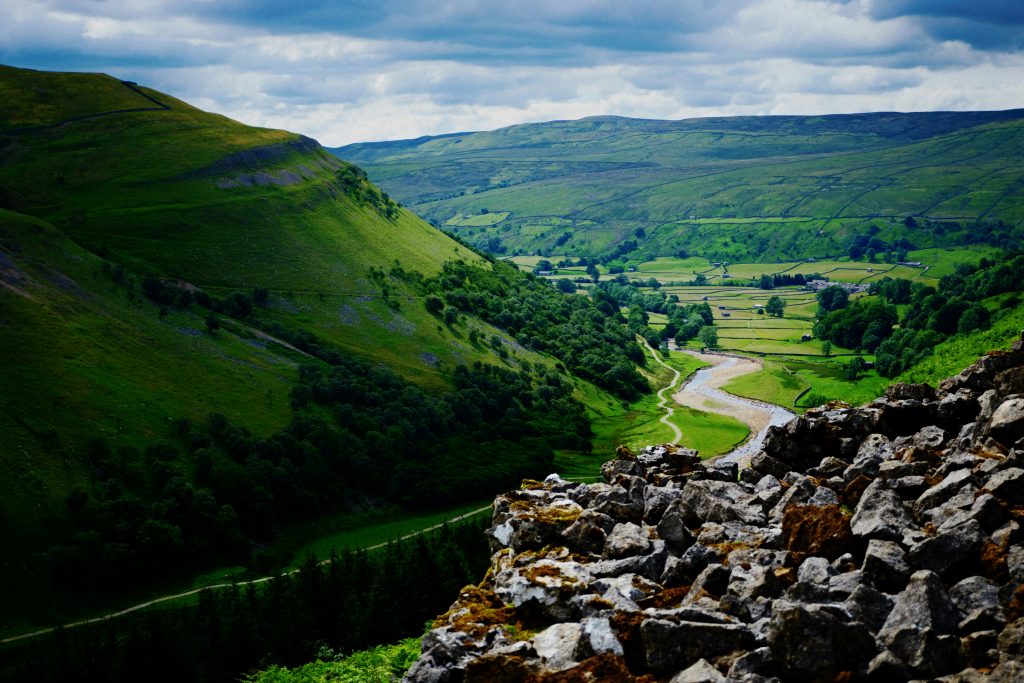 A breathtaking view of a lush green valley in Yorkshire, with rolling hills, a winding river, and traditional dry stone walls stretching across the landscape. The foreground features rugged rocks, adding depth to the scene. The sky is a mix of blue and clouds, casting soft shadows over the countryside.