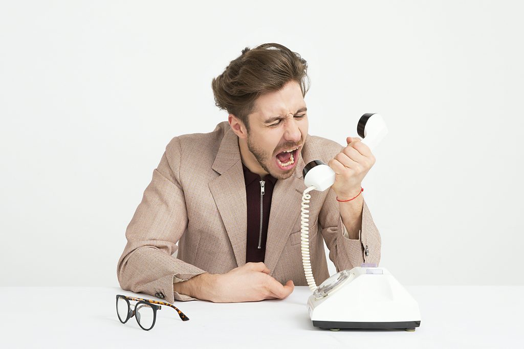 Frustrated man in a beige suit yelling into a vintage rotary phone, expressing anger or impatience. A pair of glasses rests on the table beside him.