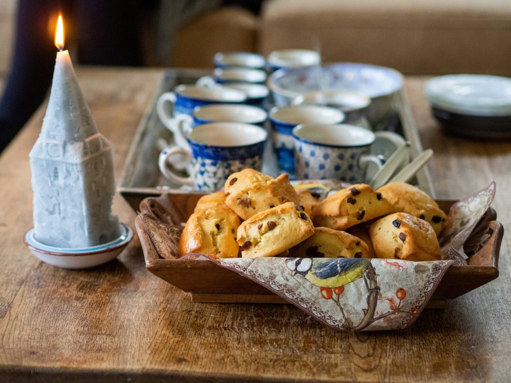 A rustic wooden table set for afternoon tea with a wooden tray of golden-brown scones studded with raisins, accompanied by blue and white patterned teacups and a lit decorative candle. The cozy setting suggests a warm and inviting British tea-time atmosphere.