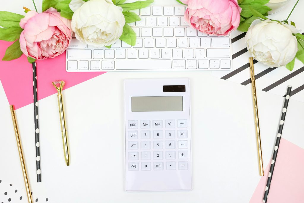 Flat lay of a white calculator on a white desk surrounded by a white keyboard, pink and white peonies, gold pens, and black-and-white polka dot straws, creating a stylish and feminine budgeting or workspace theme.