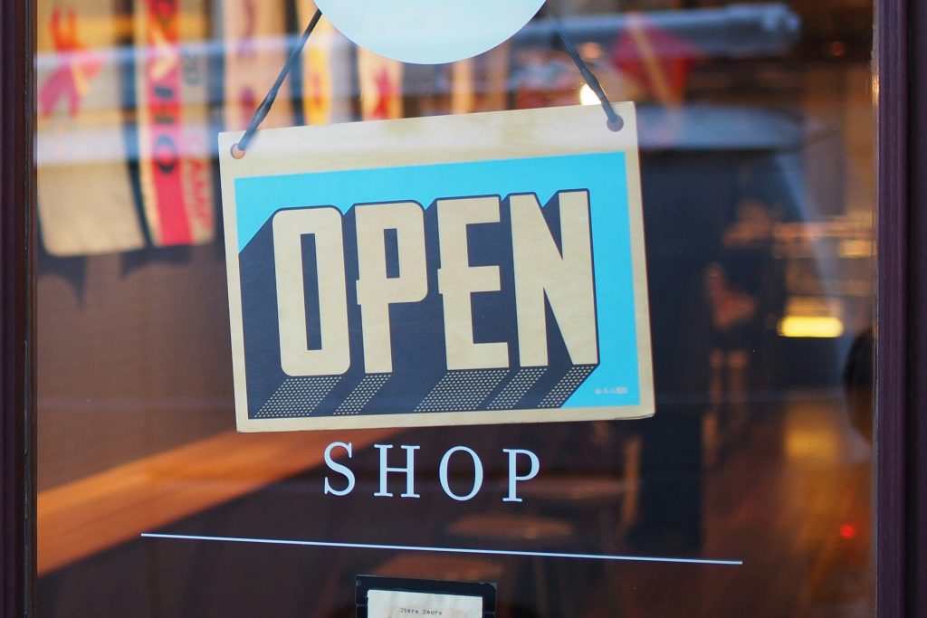 A close-up of a glass shop window displaying a wooden "OPEN" sign with bold typography and a blue background. The reflection in the glass shows a blurred interior with warm lighting and customers inside.