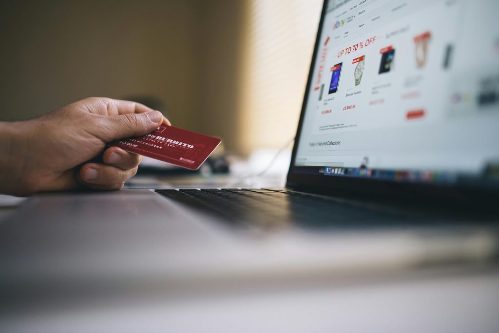 Person holding a red credit card while shopping online on a laptop, with a website showing discounts of up to 70% off.