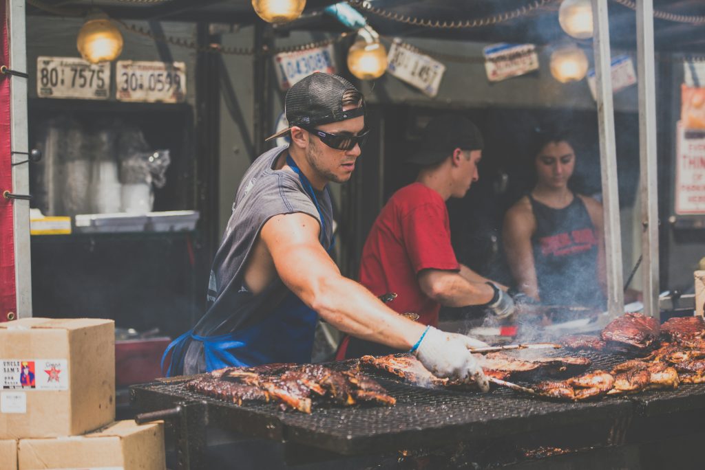 A food vendor wearing a sleeveless shirt, apron, and sunglasses grills an assortment of meats at a bustling food festival. Smoke rises from the sizzling barbecue, creating a dynamic scene. Two other team members are visible in the background, working in the food stall decorated with vintage license plates and warm hanging lights.