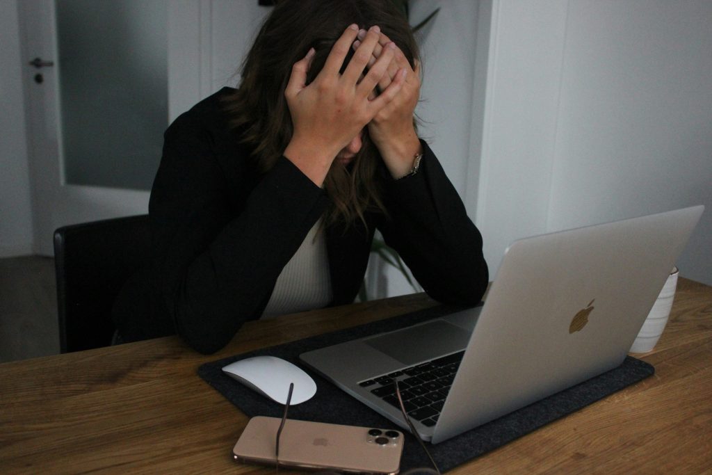 A frustrated woman sitting at a desk with her head in her hands, looking at a laptop screen. A smartphone and a pair of glasses are placed beside her on the wooden table, suggesting distress or disappointment, possibly related to online activities.