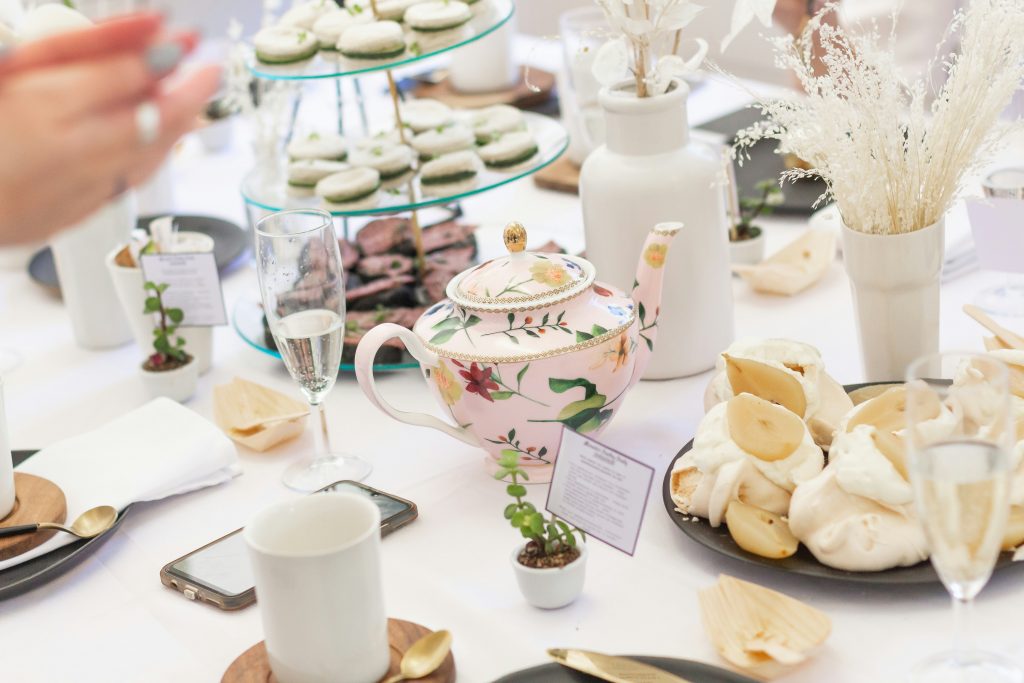 A beautifully set tea party table featuring a floral pink teapot, tiered trays with macarons and chocolate treats, elegant white tableware, and delicate dried floral arrangements. The setting exudes charm and sophistication, with guests' hands visible in the background, adding a lively touch to the scene.