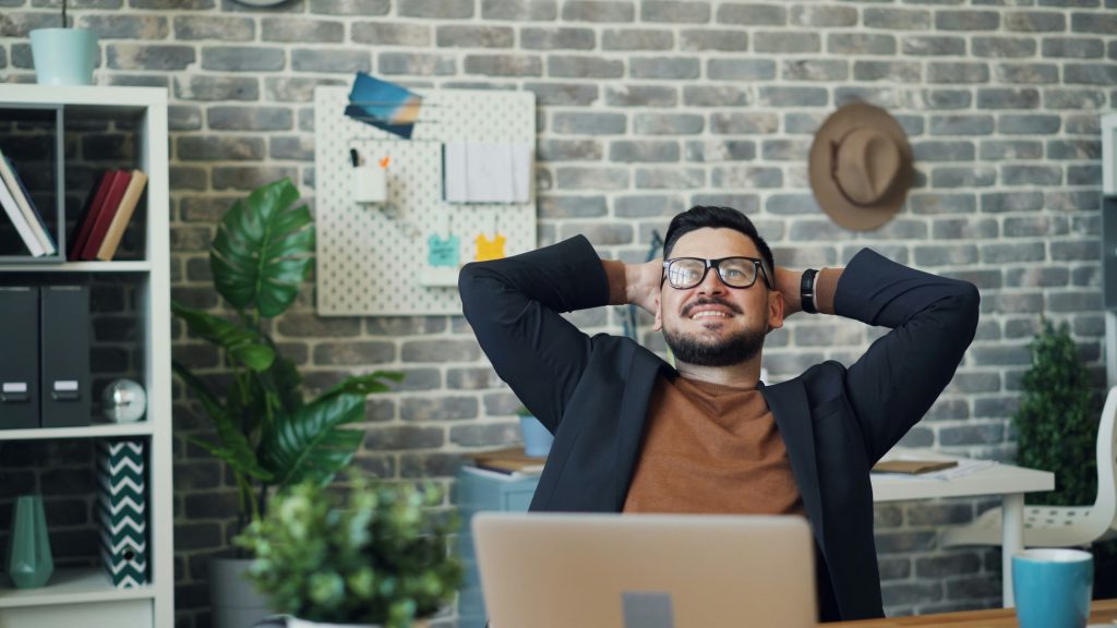 A relaxed man smiling with his hands behind his head, sitting at a desk with a laptop in a stylish home office, symbolising calmness, mindfulness, and contentment.