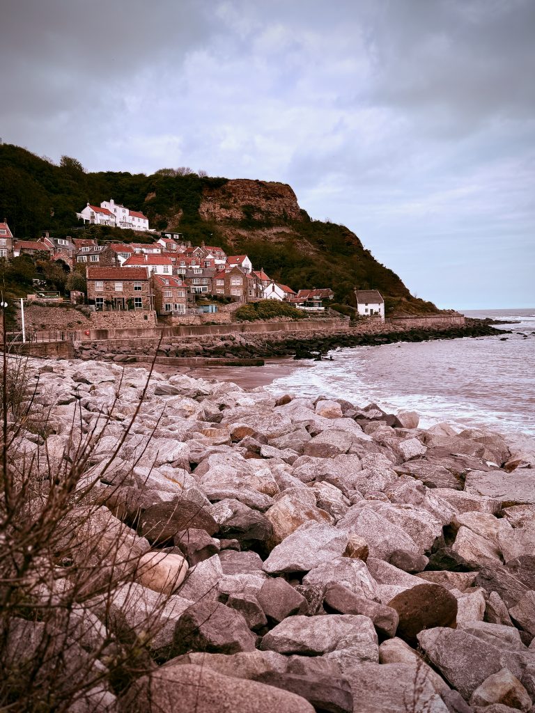 Coastal village of Runswick Bay with red-roofed cottages nestled against a cliff, viewed from a rocky shoreline on a cloudy day in North Yorkshire