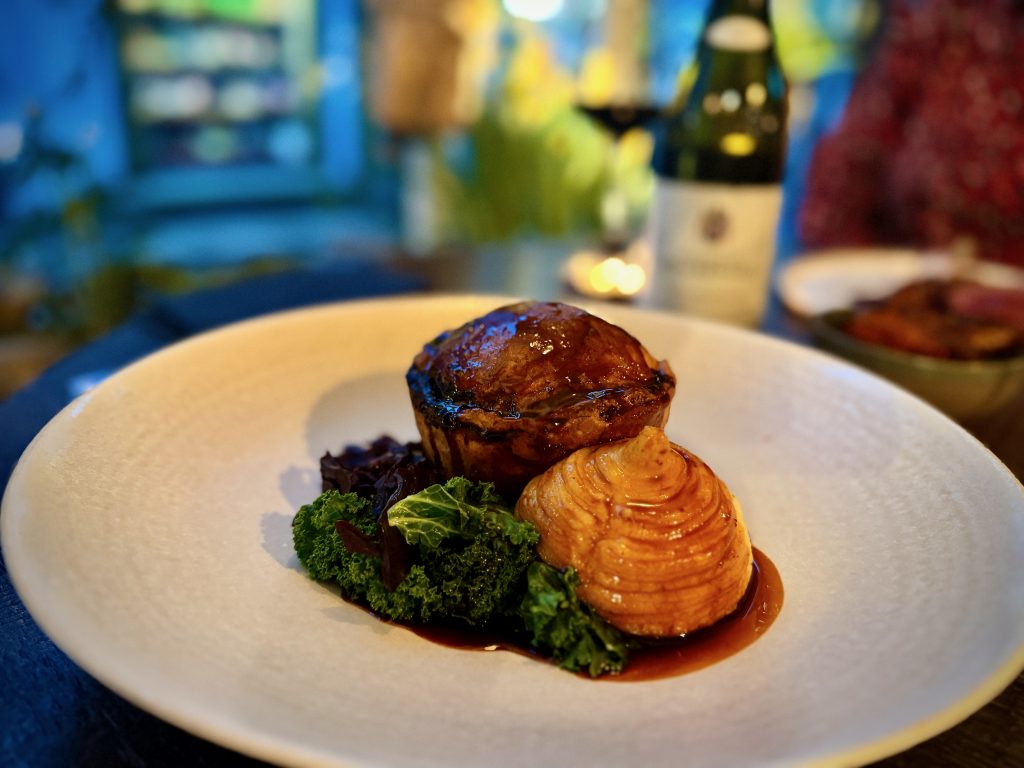 Gourmet beef pie served with layered dauphinoise potato, kale, and rich jus at The Black Bull in Sedbergh, with wine and candlelight in the background.