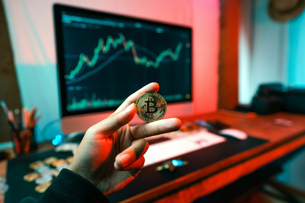 A person holds a physical Bitcoin coin in front of a computer monitor displaying a cryptocurrency trading chart. The workspace is lit with red and teal lighting, creating a modern, tech-focused atmosphere.